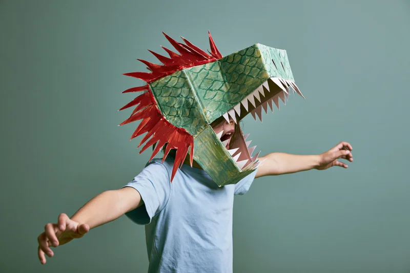Child wearing handmade cardboard dinosaur head costume with arms outstretched in studio
