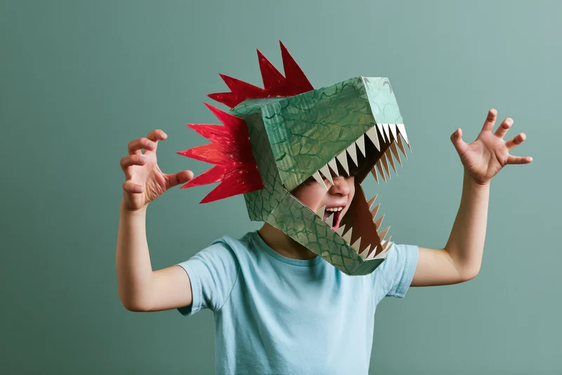 Child wearing handmade cardboard dinosaur head costume with arms raised and open mouth against plain background