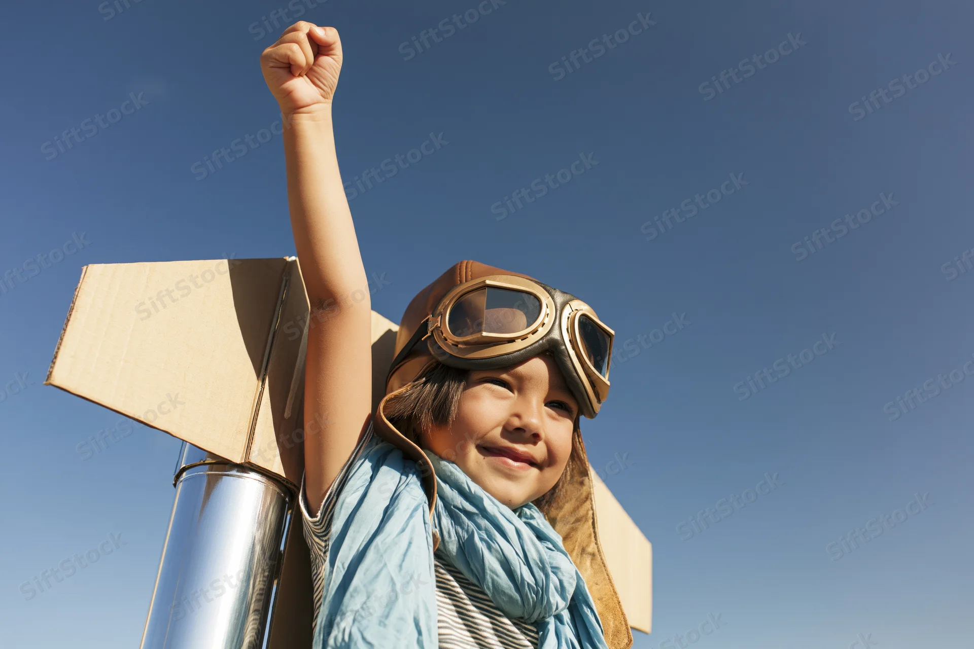 Child wearing aviator goggles and cardboard jetpack, smiling with raised arm against blue sky