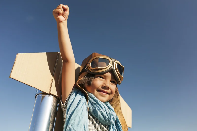 Child wearing aviator goggles and cardboard jetpack, smiling with raised arm against blue sky
