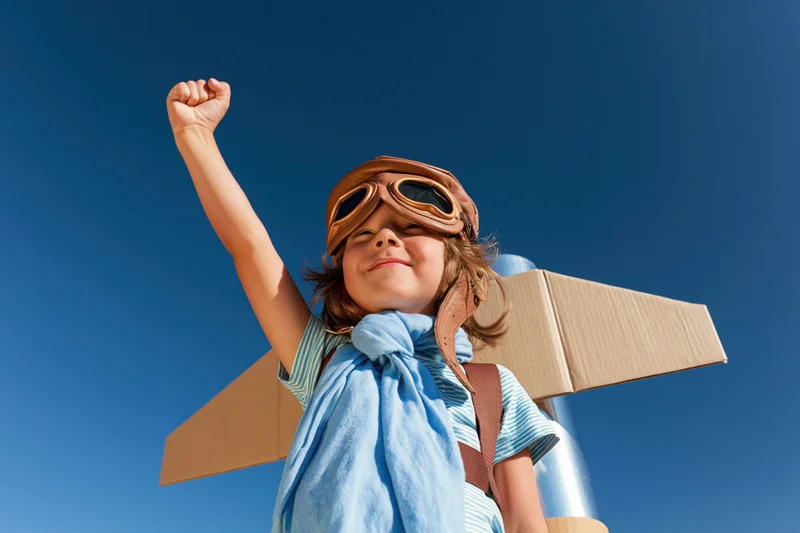 Child in aviator costume with cardboard wings raised arm against blue sky
