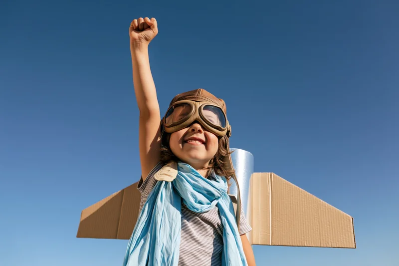 Child wearing aviator goggles and cardboard wings, raising arm against blue sky