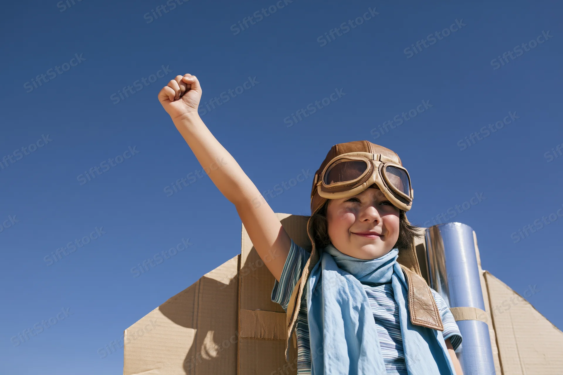 Child in aviator costume with cardboard rocket, smiling and raising fist against blue sky