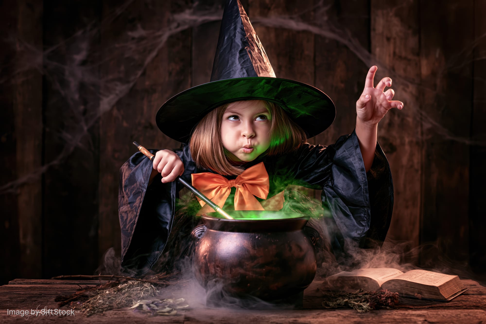 Young girl in witch costume stirring glowing green potion in cauldron at wooden table with open book and cobwebs