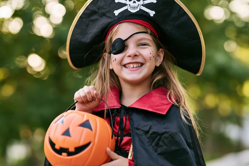 Young girl in pirate costume smiling while holding orange pumpkin bucket outdoors