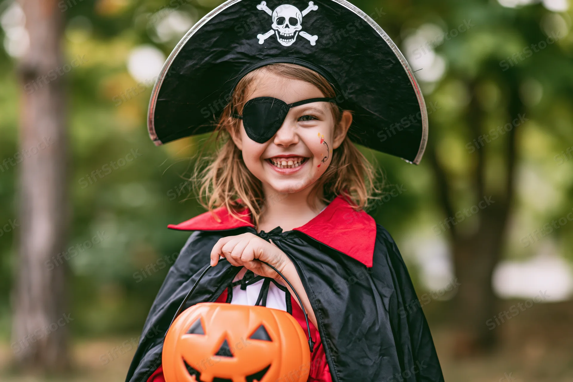 Young girl in pirate costume smiling while holding orange jack-o-lantern bucket outdoors
