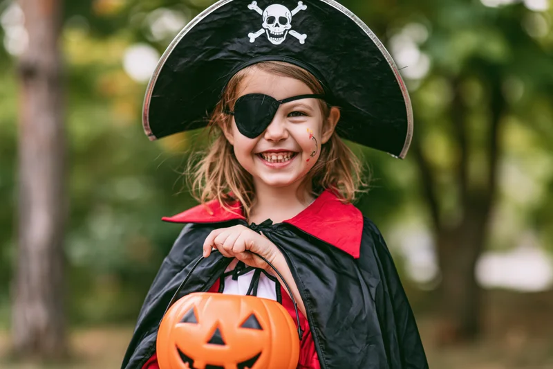 Young girl in pirate costume smiling while holding orange jack-o-lantern bucket outdoors