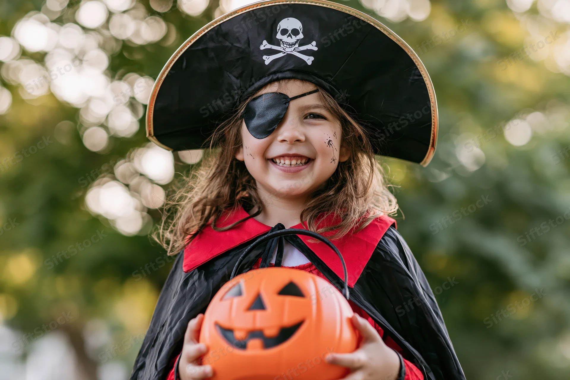 Young girl in pirate costume smiling while holding orange jack-o-lantern bucket outdoors