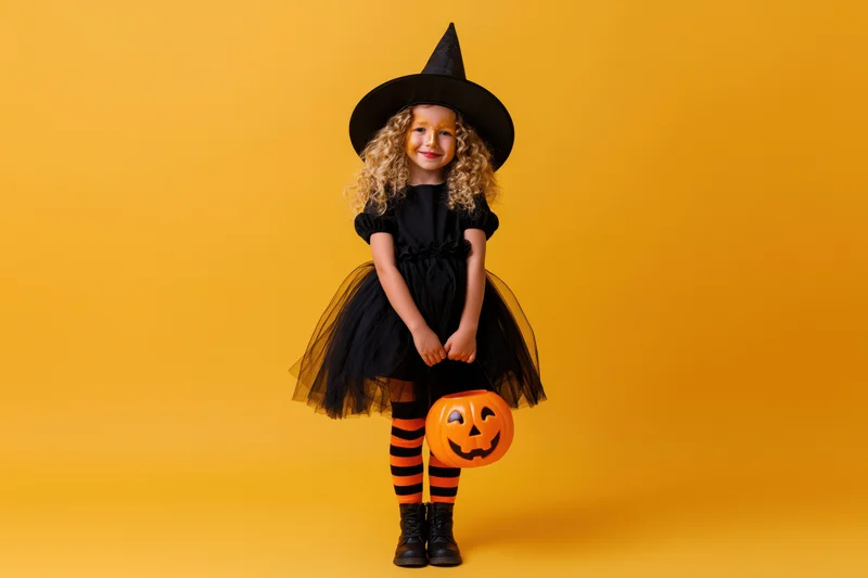 Young girl in witch costume holding pumpkin bucket against orange background