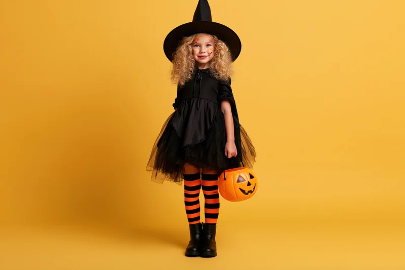 Young girl in witch costume holding pumpkin bucket against yellow background