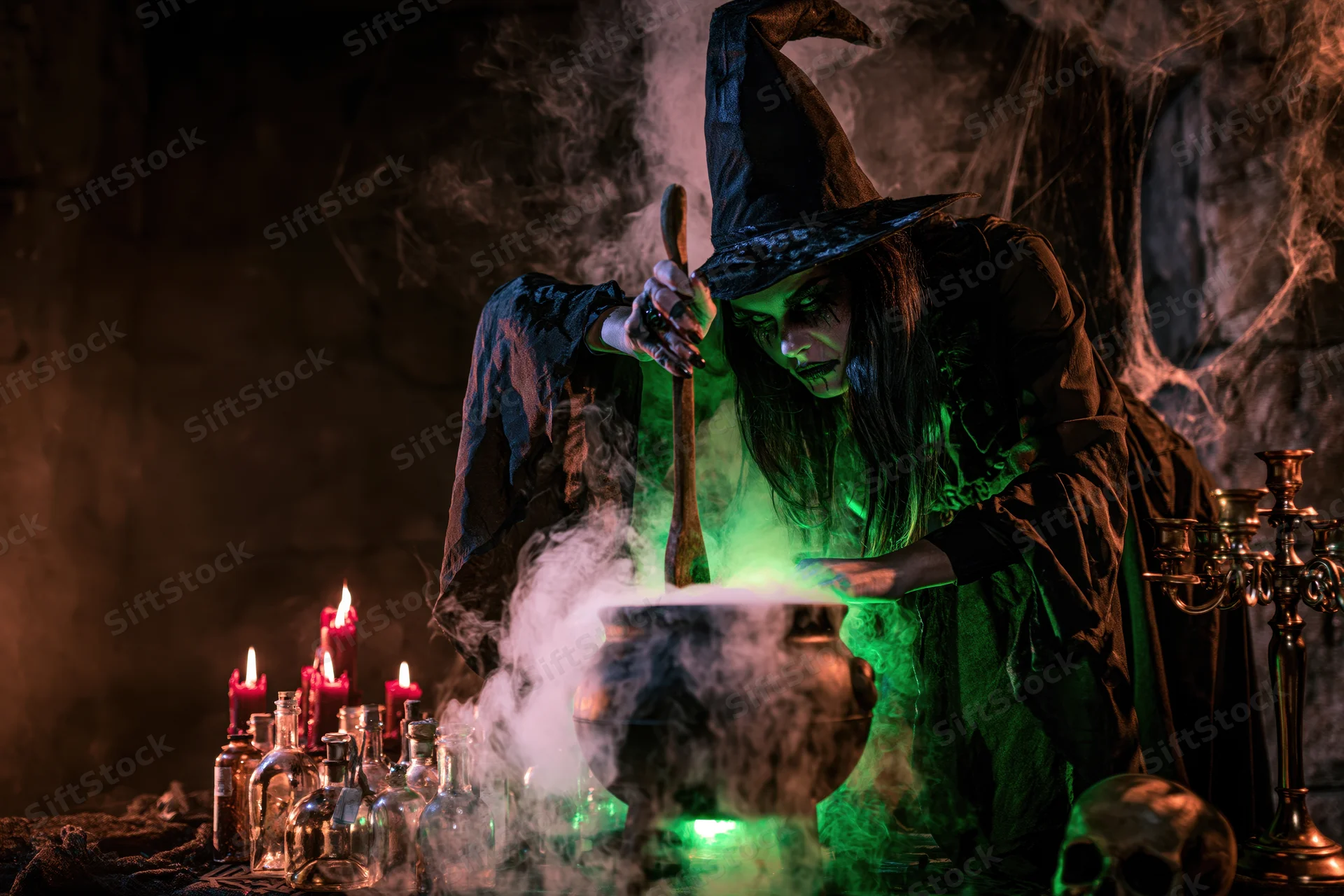 Witch in costume stirring a glowing cauldron in a dark cave with candles and potion bottles around