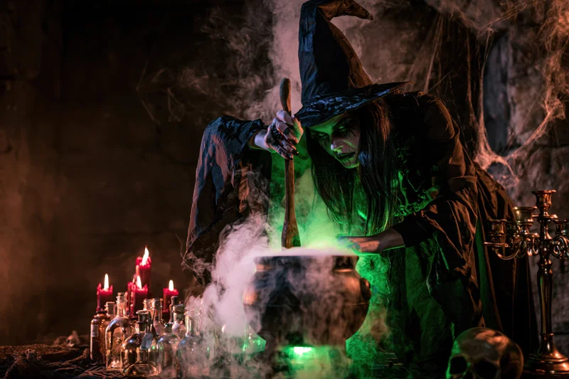 Witch in costume stirring a glowing cauldron in a dark cave with candles and potion bottles around