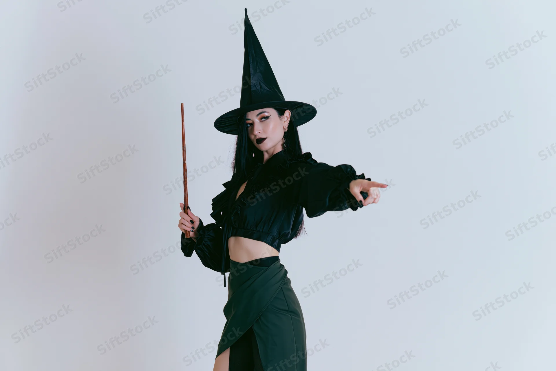 Woman in witch costume holding wooden wand and pointing against white background