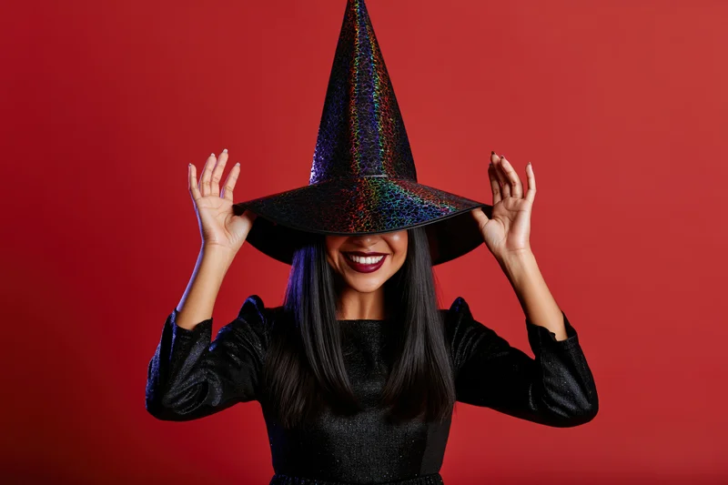 Woman in witch costume holding hat against red background smiling