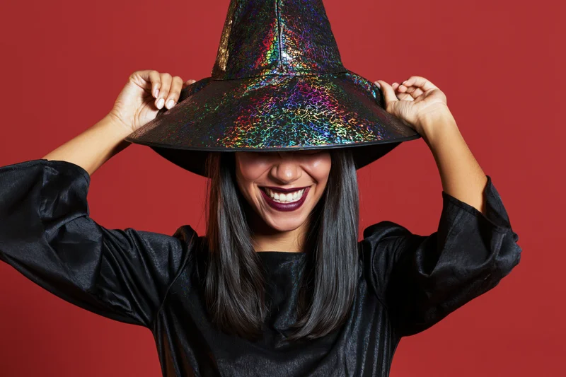 Woman in witch costume tilting hat against red background