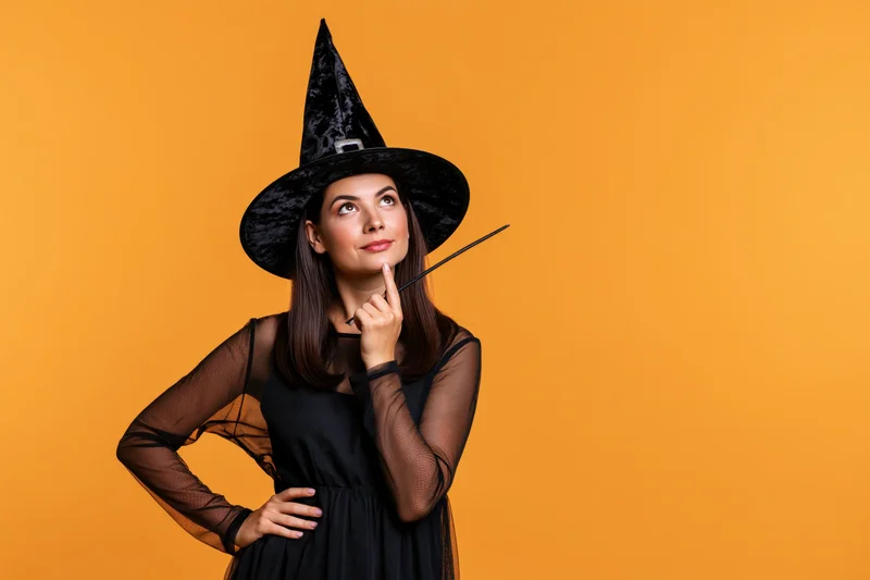Woman in black witch costume pointing finger to chin against orange background