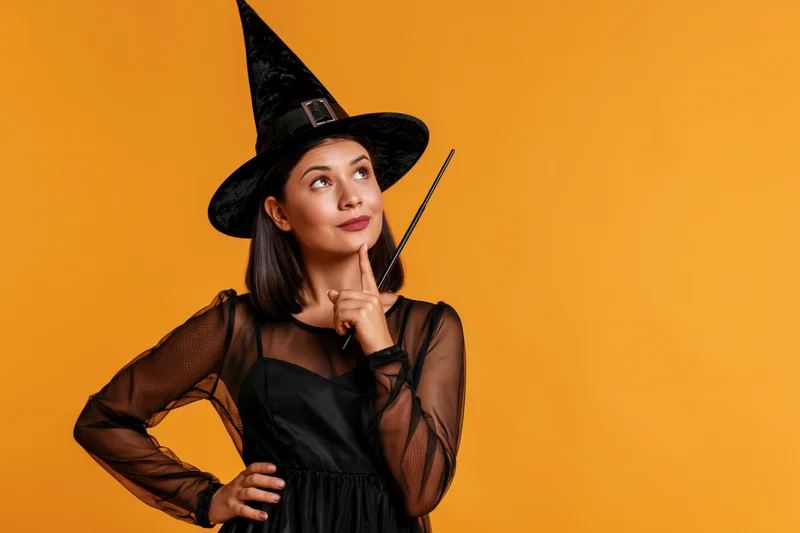 Woman in black witch costume holding wand and pointing finger to chin against orange background