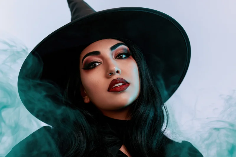Woman in wide-brim black witch hat with dramatic makeup and smoke swirling around her face