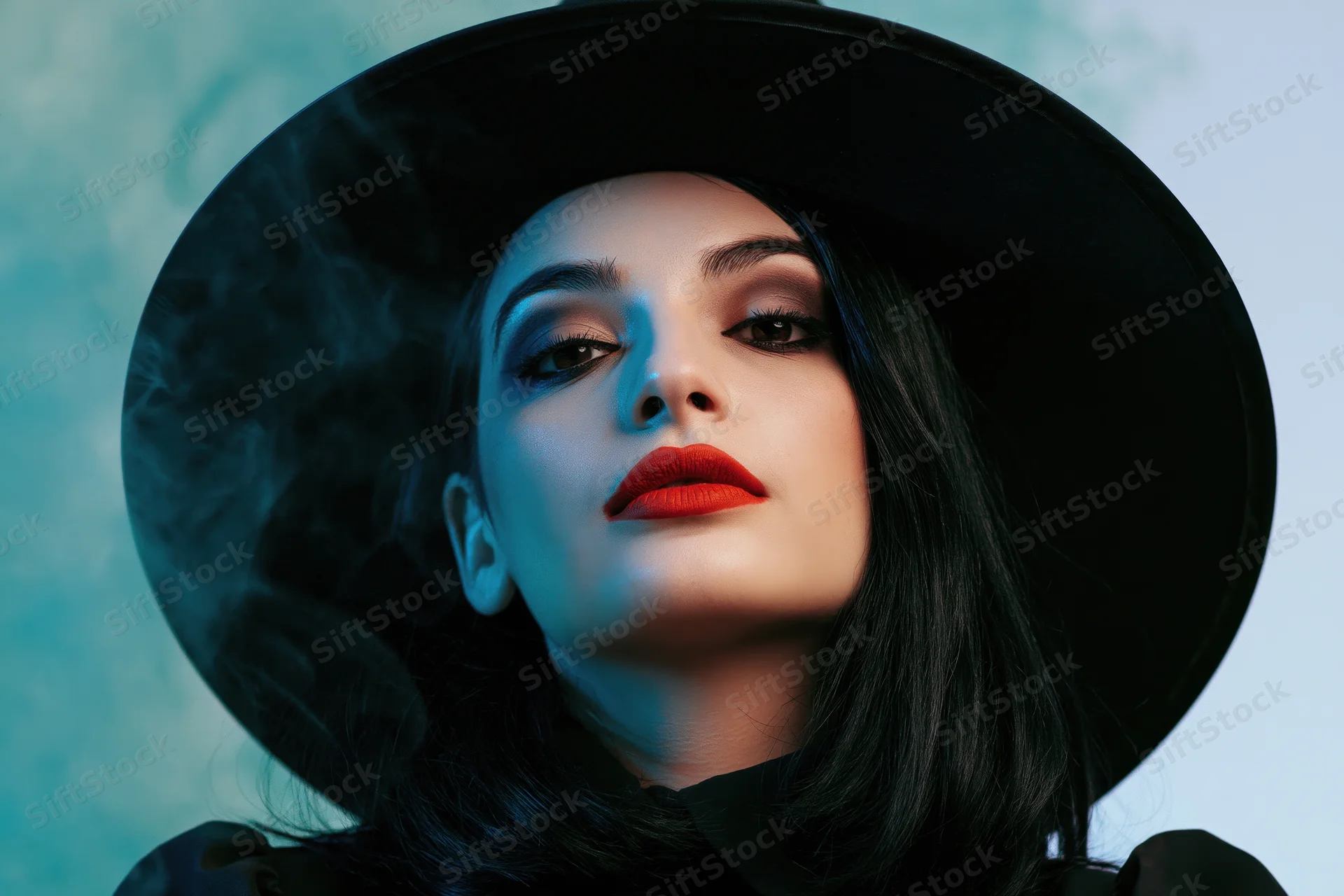 Young woman in wide-brim black witch hat looking directly at camera with dramatic makeup and red lips in studio setting