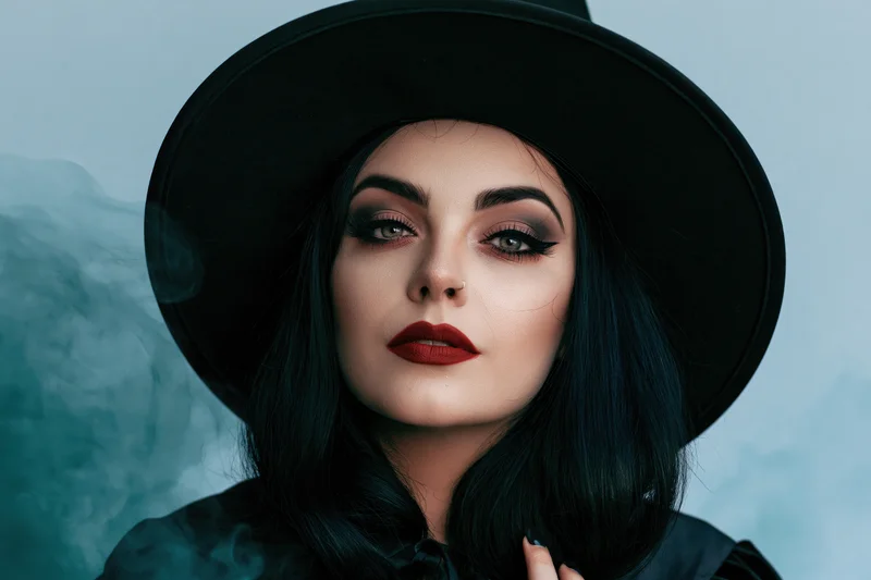Woman in wide-brim black witch hat with dark hair and red lips, looking directly at camera against smoky blue background