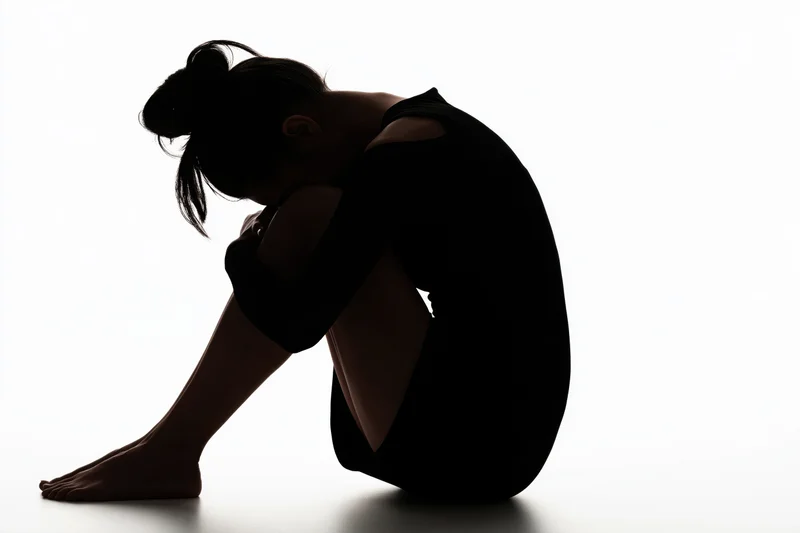 Silhouette of a young woman sitting on the floor with knees pulled to chest and head down