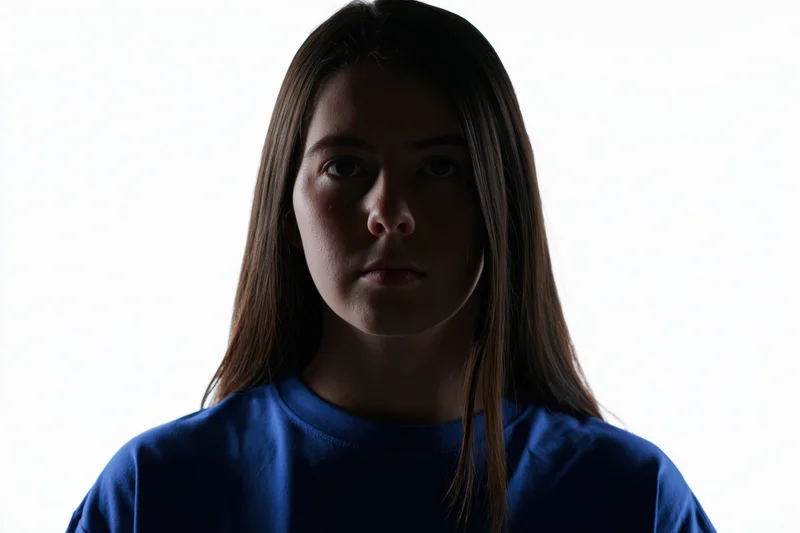 Young woman with long straight hair facing camera in studio lighting, wearing blue tshirt against white background