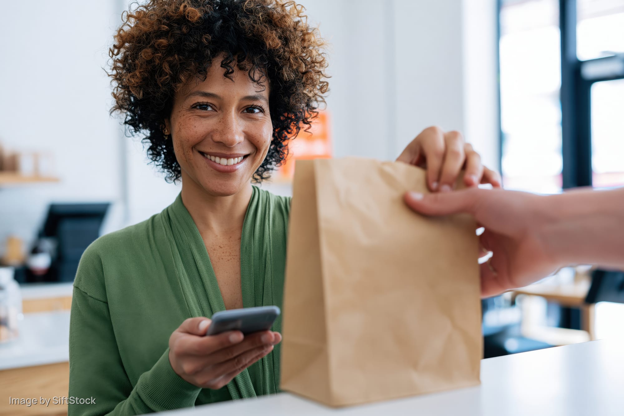 Woman smiling while receiving a paper bag at a store counter, holding a smartphone