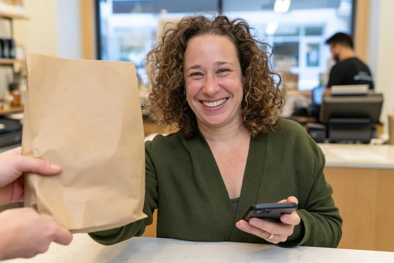 Woman smiling at counter while receiving paper bag and holding phone