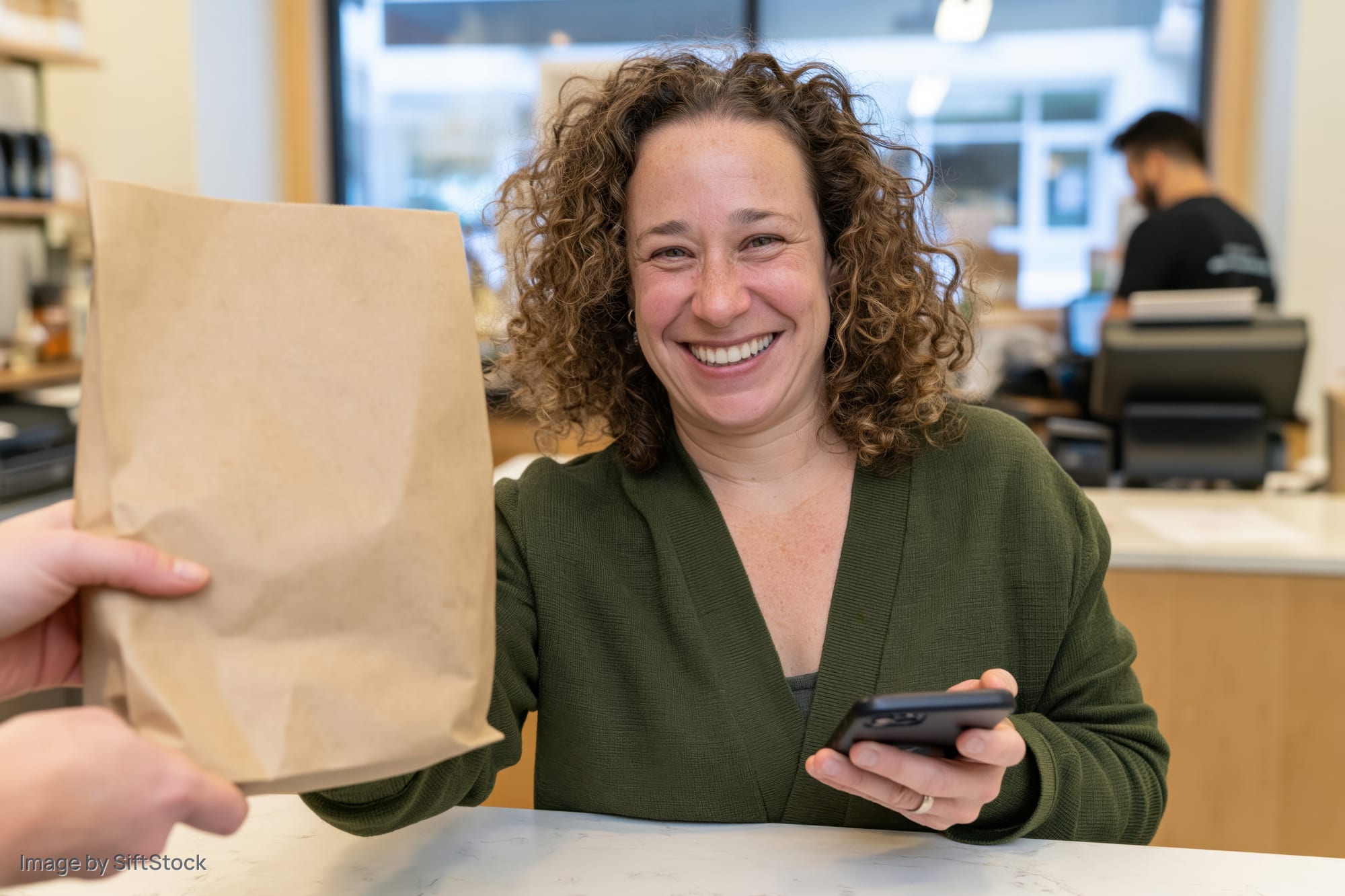 Woman smiling at counter while receiving paper bag and holding phone