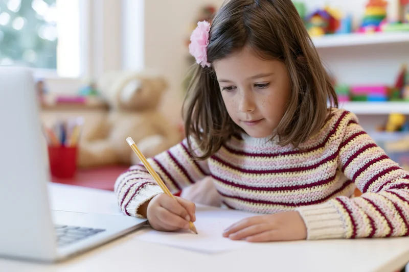 Girl writing with pencil at desk beside laptop in home study area