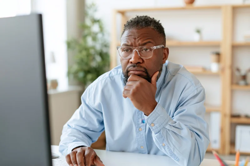 Man in light blue shirt and clear glasses concentrating at desk in front of computer monitor