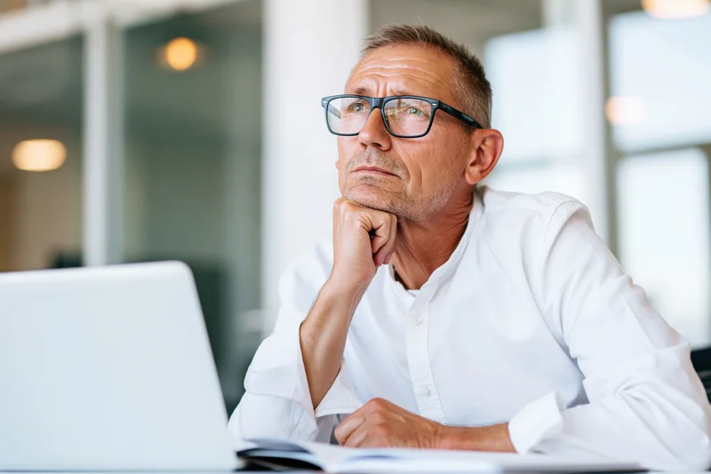 Man in white shirt thinking at desk with laptop in office
