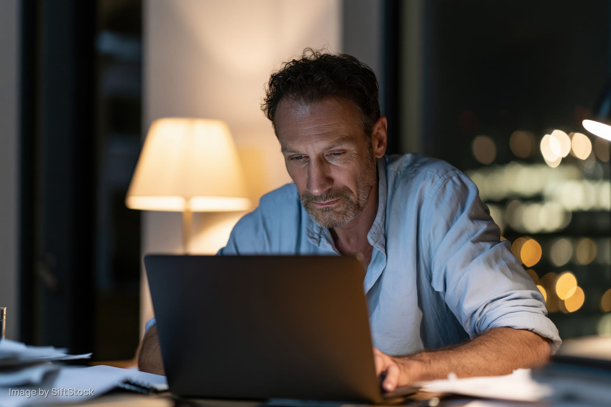 Man typing on laptop at cluttered desk at night by window with city lights visible outside