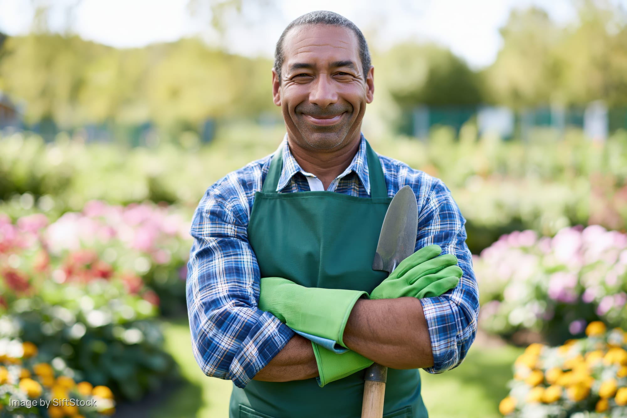 Smiling man in green apron holding trowel with arms crossed in garden