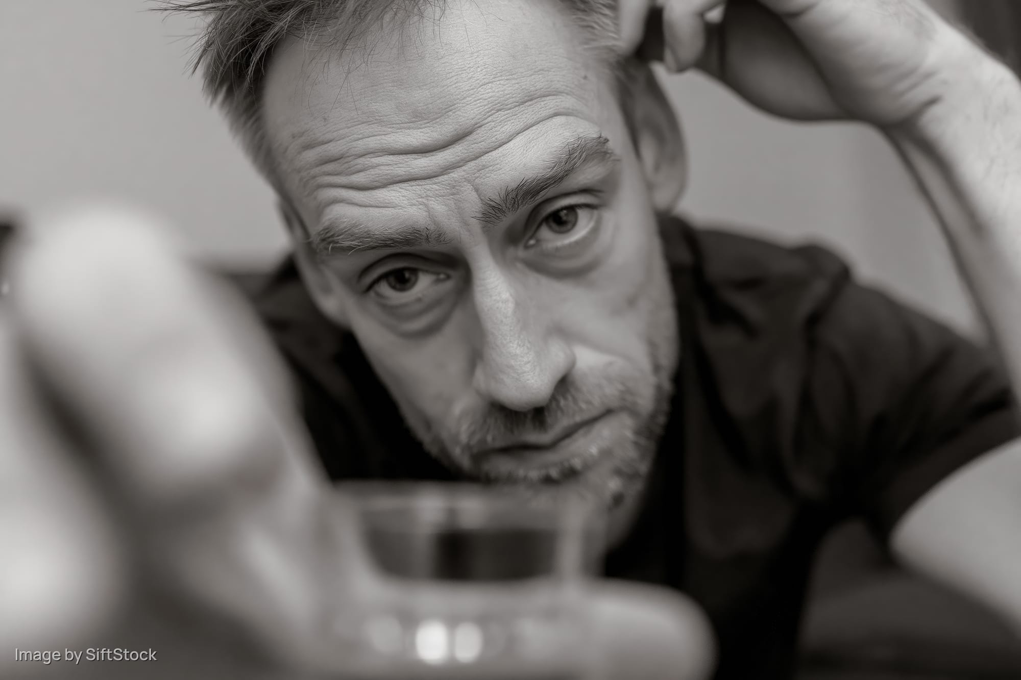 Man with short hair and stubble leaning forward, eyes downcast, hand on head, holding glass in foreground
