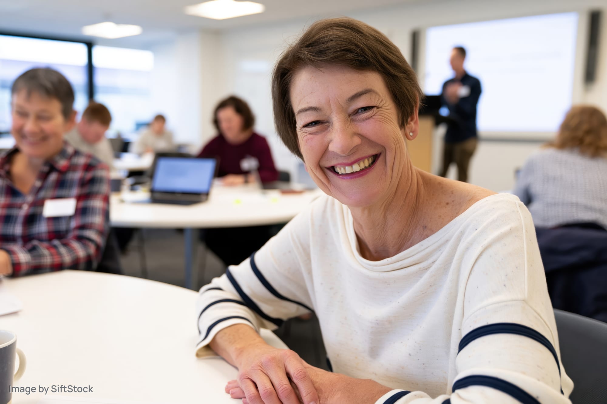 Smiling woman sitting at table in workshop with participants and instructor presenting on screen