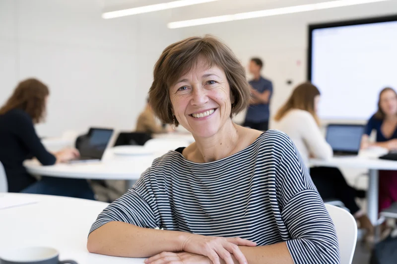 Middle-aged woman smiling at camera in office meeting with colleagues at tables using laptops