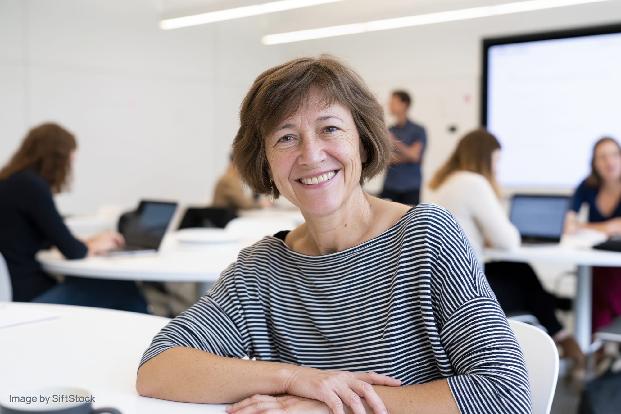 Middle-aged woman smiling at camera in office meeting with colleagues at tables using laptops