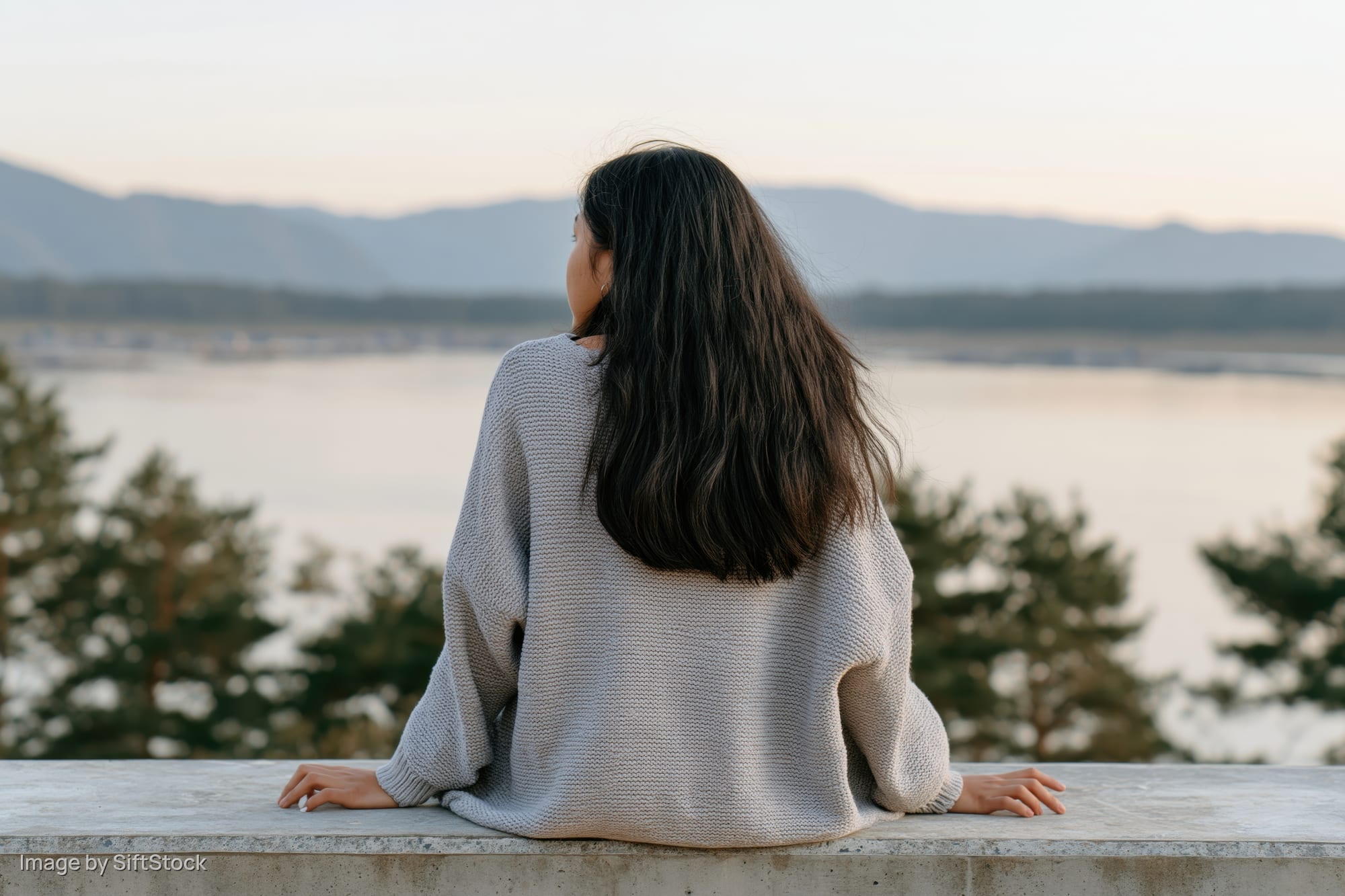 Woman in gray knit sweater sitting on balcony overlooking lake and mountains at dusk