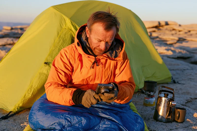 Man in orange jacket sitting on blue sleeping bag beside green tent, reviewing camera in rocky outdoor setting at sunrise