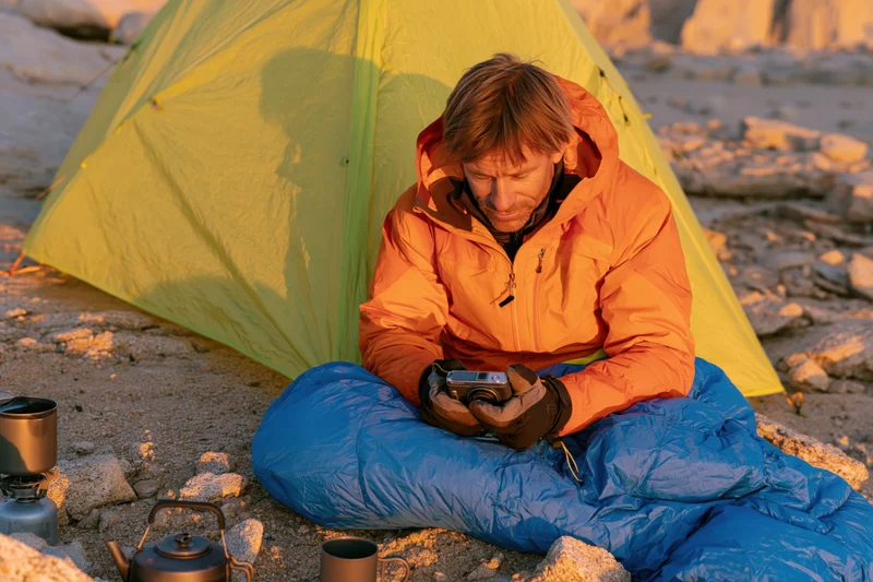 Man in orange jacket sitting in blue sleeping bag reviewing camera near yellow tent in rocky mountain terrain at sunrise