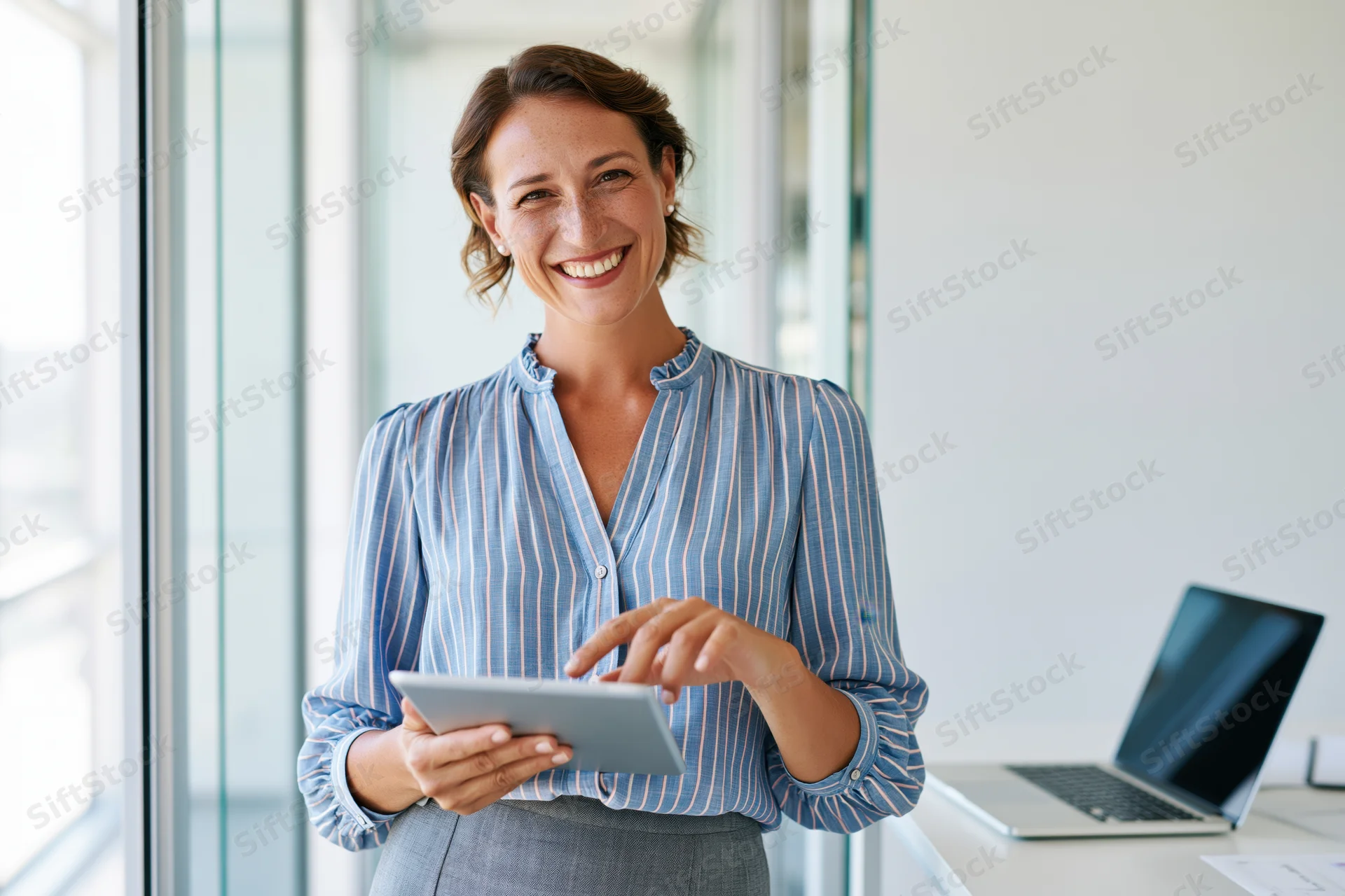 Smiling woman holding tablet in bright modern office with laptop on desk