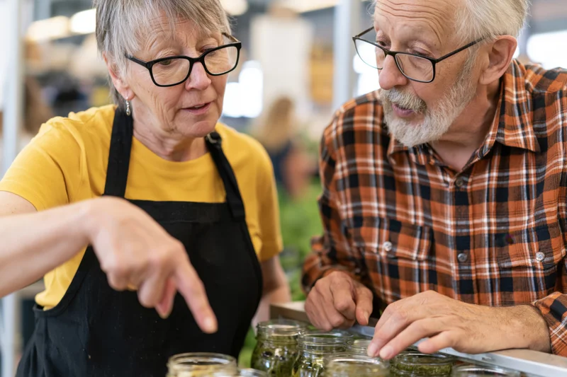 Senior woman vendor in yellow shirt and black apron pointing at jars of preserved food while discussing with elderly man in plaid shirt at market display