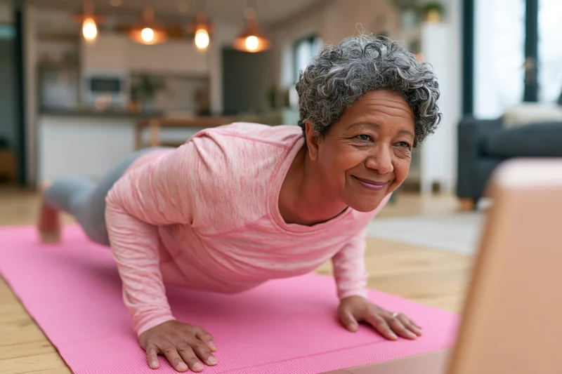 Senior woman doing plank exercise on pink mat in living room while smiling at laptop screen
