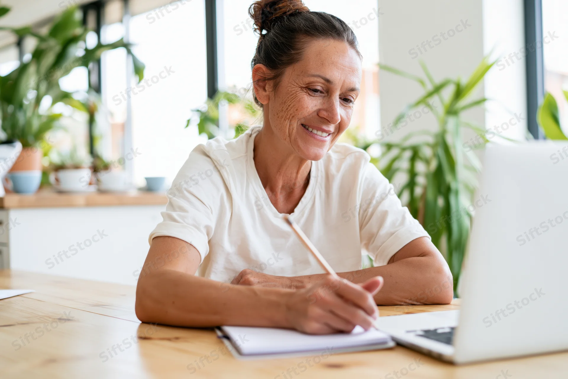 Woman smiling while writing in notebook at desk with laptop and plants nearby
