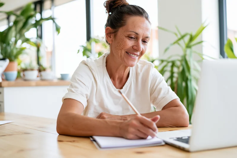 Woman smiling while writing in notebook at desk with laptop and plants nearby