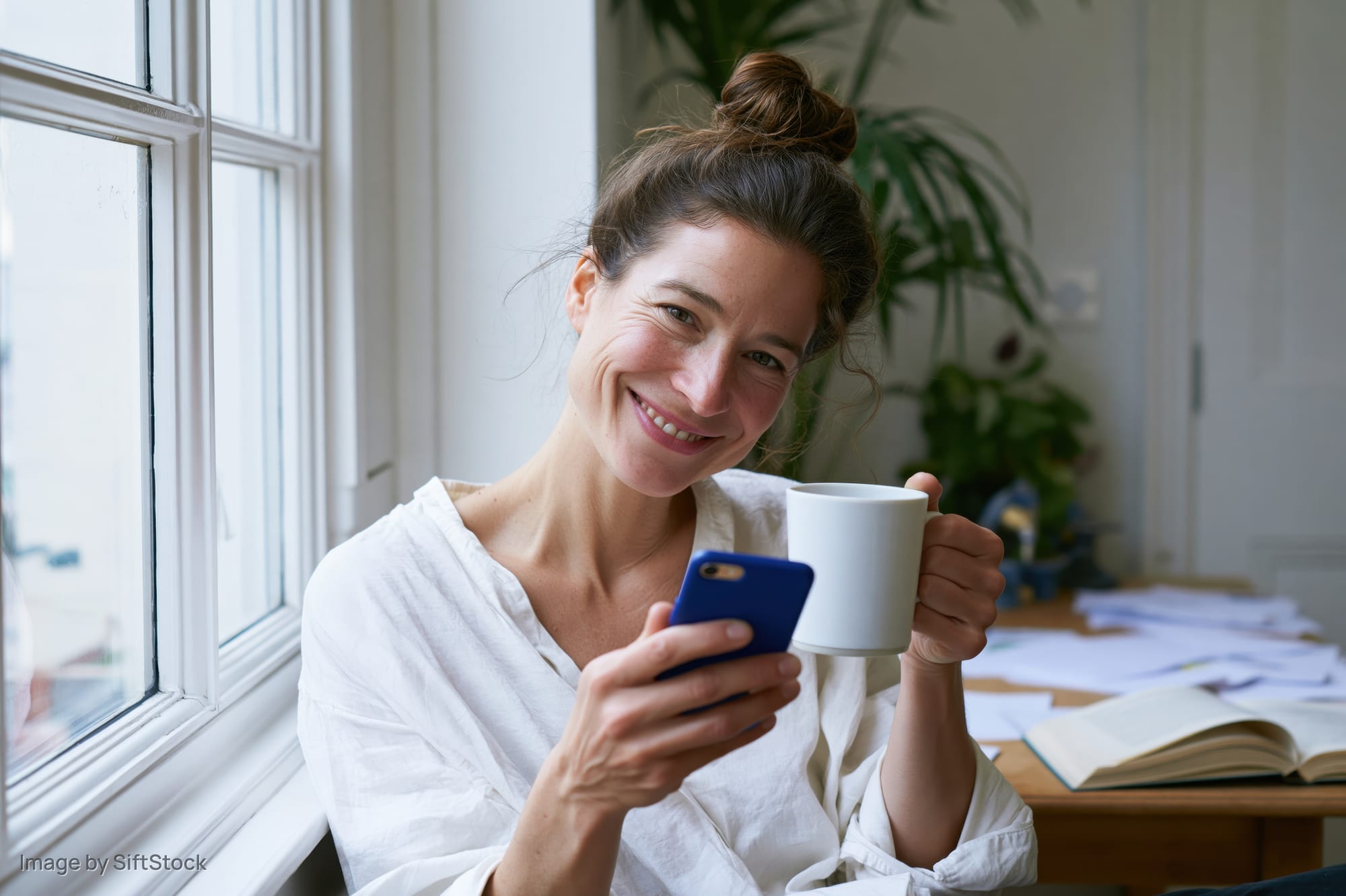 Woman smiling while holding smartphone and mug by window in home office