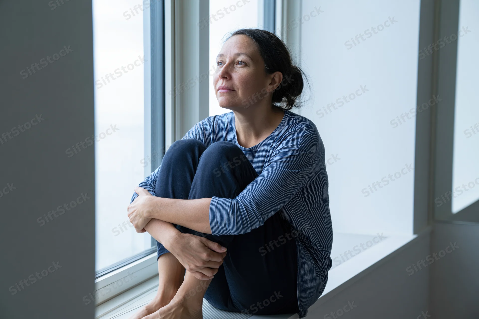 Woman sitting on windowsill with knees drawn up, looking out the window in a quiet indoor space