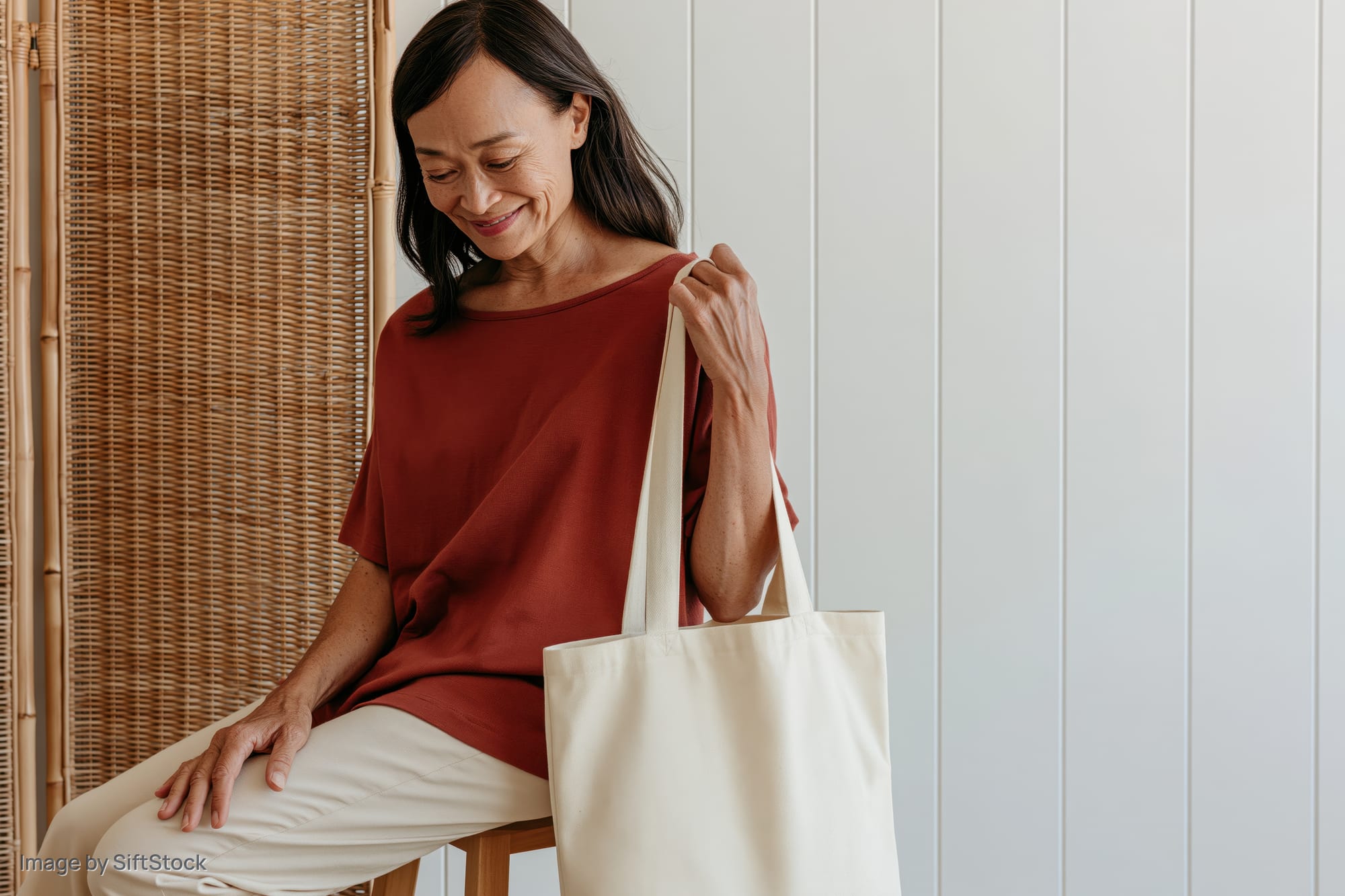 Woman sitting sideways on a wooden stool smiling softly while holding a cream tote bag