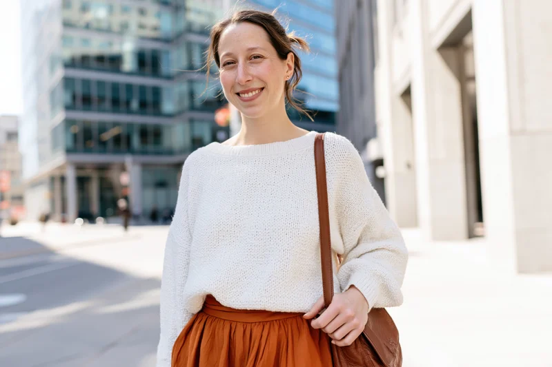 Woman smiling on city sidewalk wearing white sweater and brown skirt with leather handbag